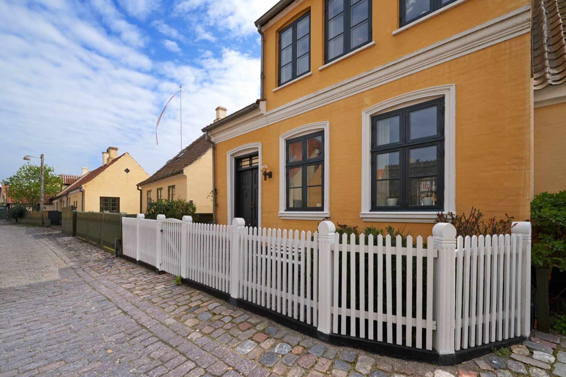 En solgul villa med mörka fönster och en vit port av trä längs en gata av kullersten