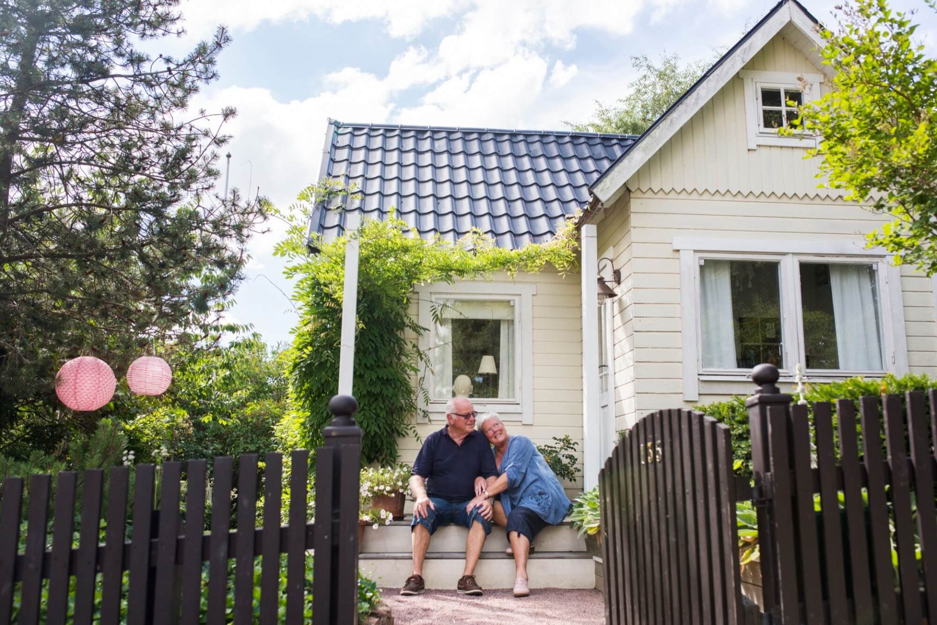 På en veranda av ett trähus målat i en varm gulfärg med svart tak, sitter ett äldre par omgivna av den djupa grönskan som omger dem